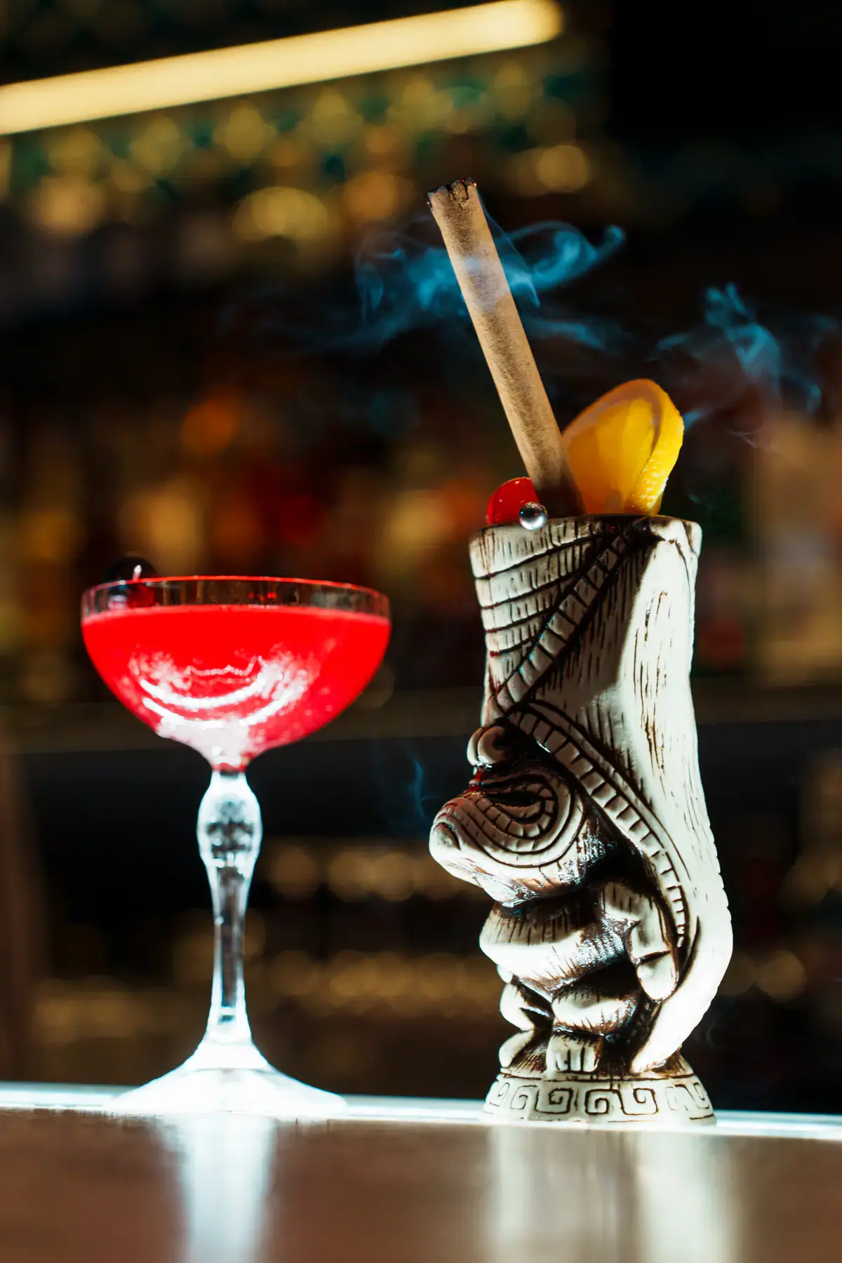 A red coupe cocktail beside a tiki mug cocktail on the bar