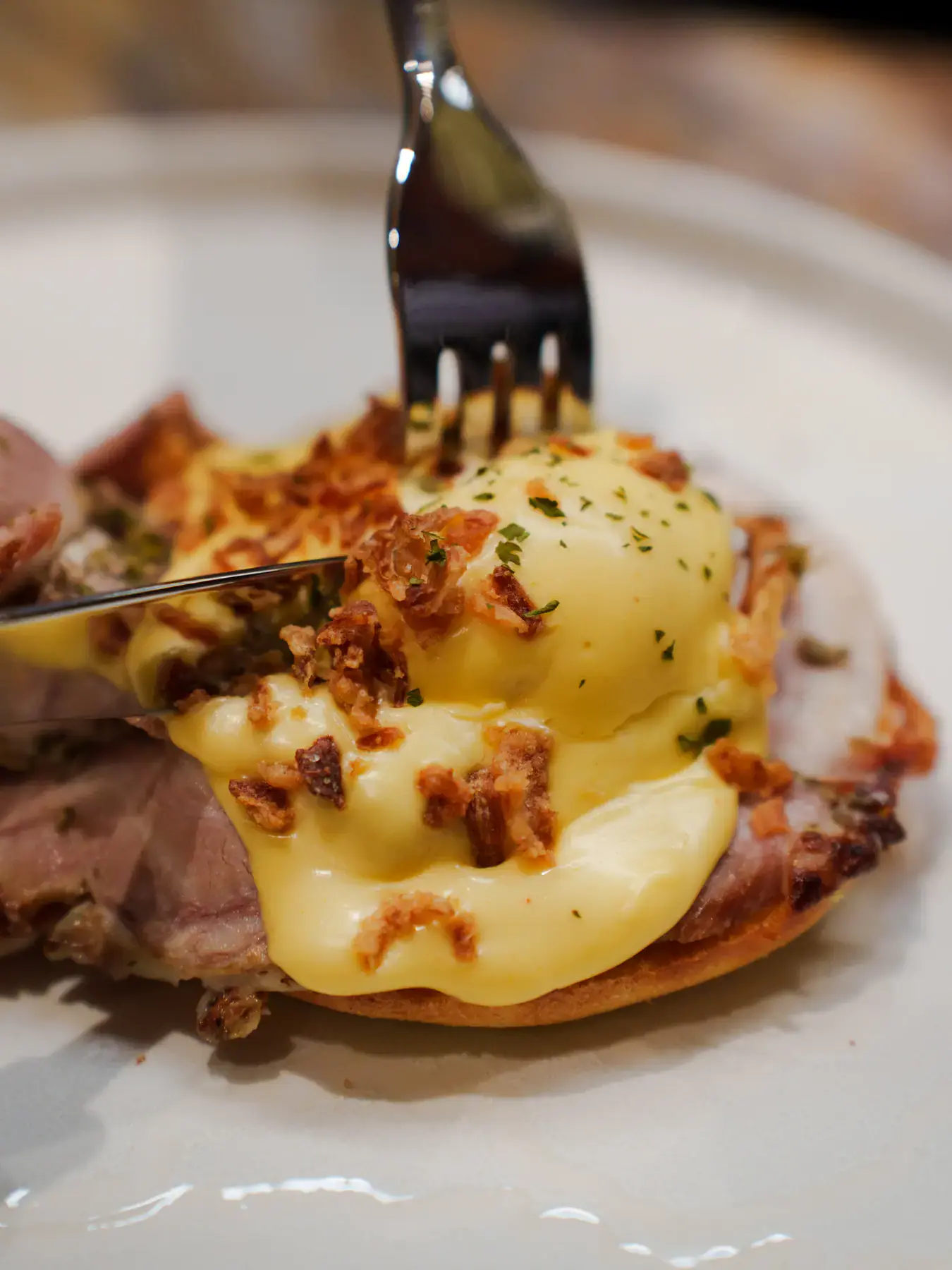 A fork cutting into a poached egg and hollandaise on an eggs Benedict